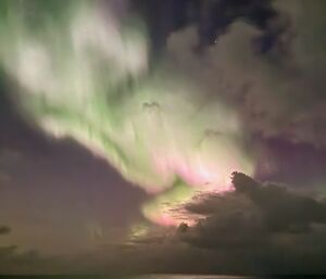A very colourful aurora with shades of white, green and pink dances in the night sky illuminating the clouds and ocean.