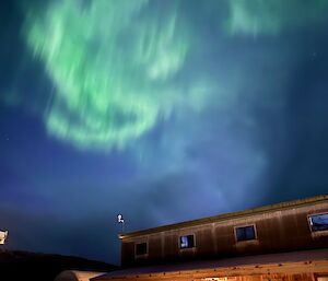 Swirling bright green lights in the night sky over a brown wood clad building with two small vehicles in the foreground.