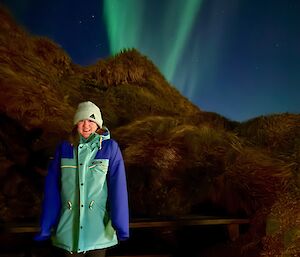 A smiling woman wearing a coat and beanie stands on the foreground with hills behind her and two bright green shards of light piercing the night sky above her.