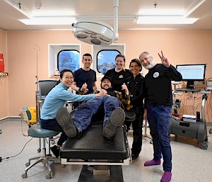 Five people stand around a bed in a medical facility. There is a person laying on the bed with their thumbs up and they are all smiling.