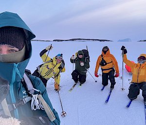 One person is taking a selfie and four people in snow gear and skis kneel behind them.