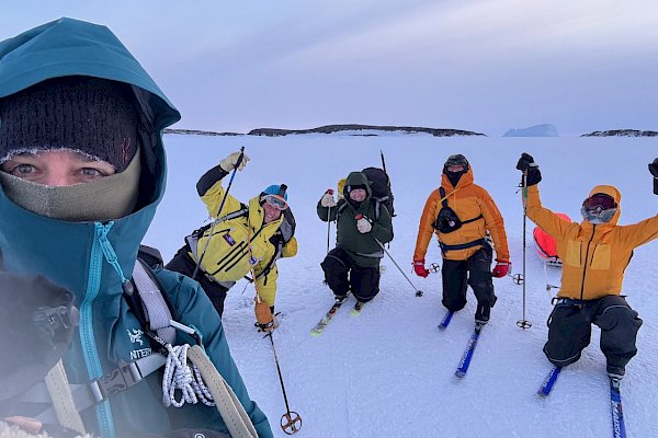 One person is taking a selfie and four people in snow gear and skis kneel behind them.