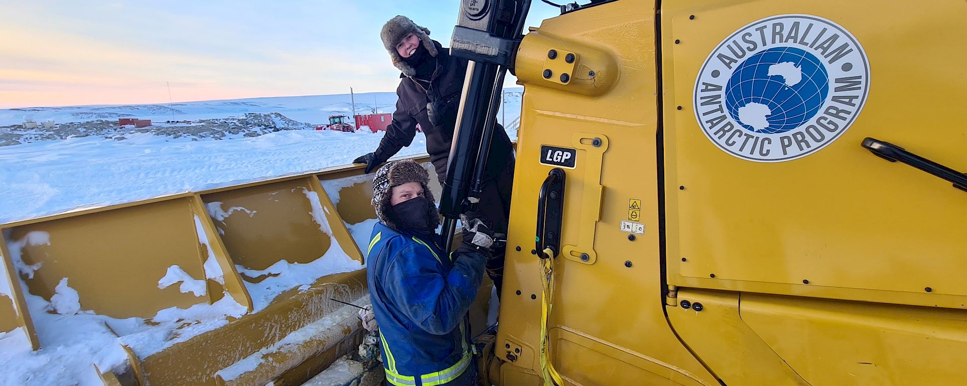 Two expeditioners dressed in cold weather gear in front of a yellow loader
