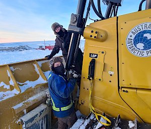Two expeditioners dressed in cold weather gear in front of a yellow loader
