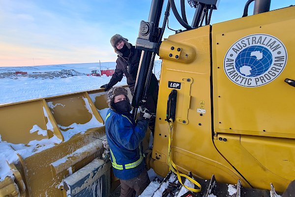 Two expeditioners dressed in cold weather gear in front of a yellow loader