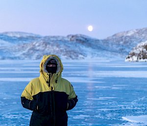 person standing on sea ice with moon and hills in background