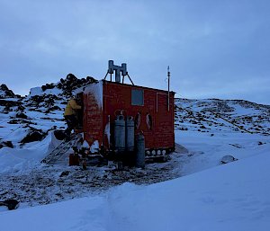 A small red hut anchored to rock and surrounded by snow.