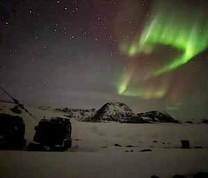 A green aurora over snow-covered, rocky mountains, with the silhouette of a hut in the foreground.