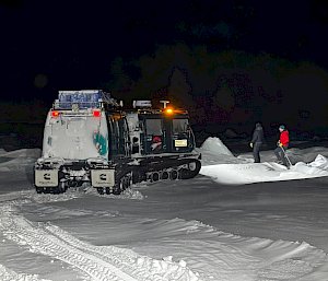 Two people measuring sea ice thickness in the light cast from the headlights of a Hagglund vehicle