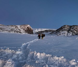 A team of four people carrying backpacks walking through deep snow.