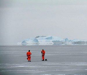 Two people in bright orange suits drilling in to sea ice