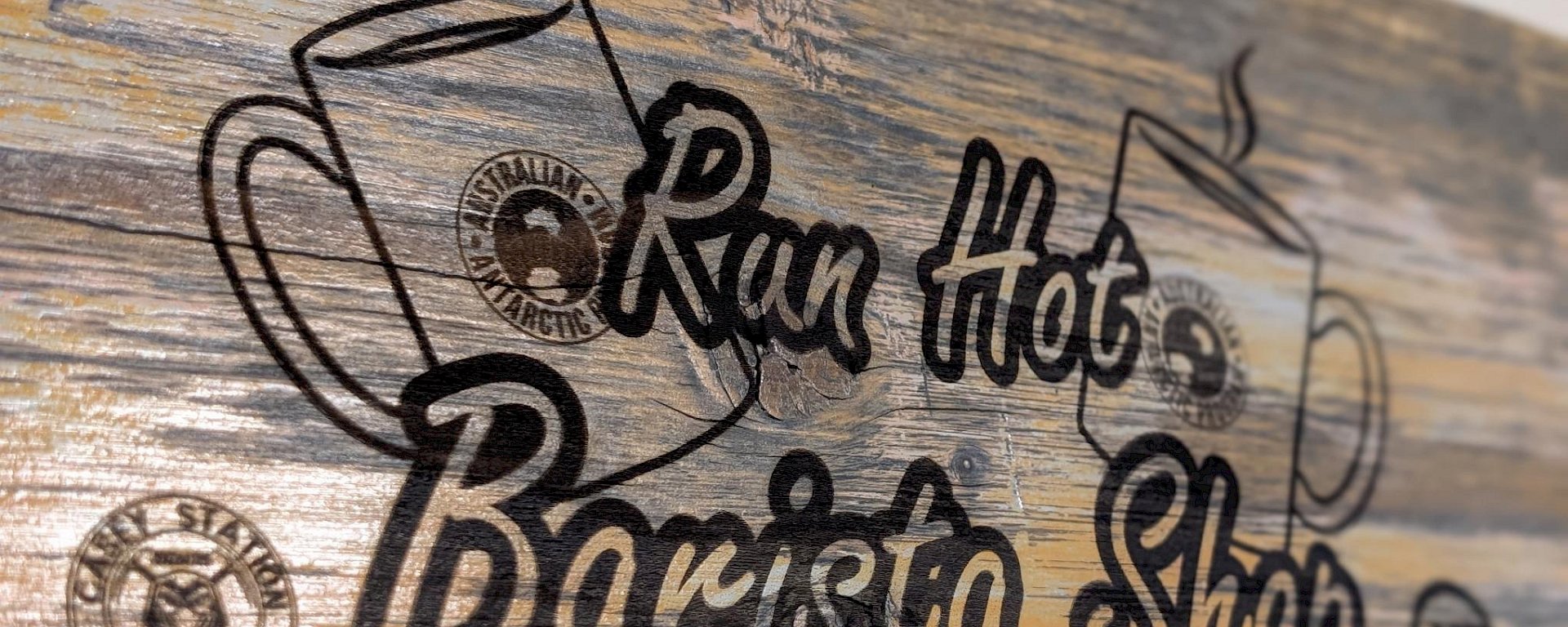 A wooden sign with Run Hot Barista Shop etched on it, alongside a picture of a mug and the station logo