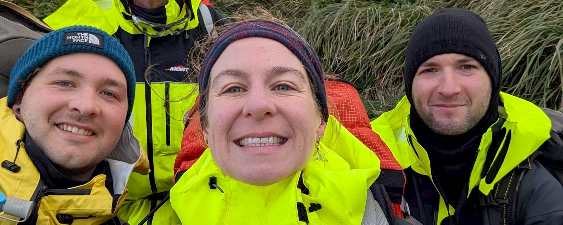 Four smiling people wearing bright yellow field gear stand amidst green tussock.