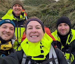 Four smiling people wearing bright yellow field gear stand amidst green tussock.