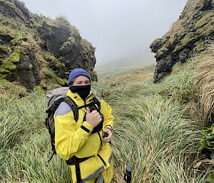 A man wearing a blue beanie and yellow outdoor gear stands with his thumb up amidst green tussock and rocky outcrops