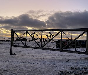 The sunset on a snow-covered beach viewed through a metal gate with the words 'Macca" emblazoned on it.