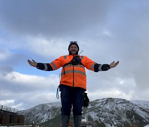 A smiling man wearing an orange jacket stands with his arms open with snow covered hills visible in the background.