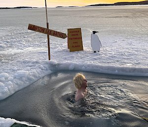 A man in a swimming hole cut in the ice with a cardboard penguin and a sign saying 'Mawson Sea Baths'