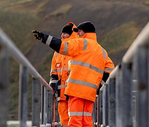 Two men in bright orange safety wear stand on a metal bridge pointing into the distance.