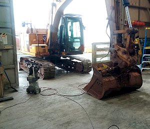 A yellow telehandler with tracks instead of wheels is half inside a large, shed opening. There is equipment on the ground to service it.