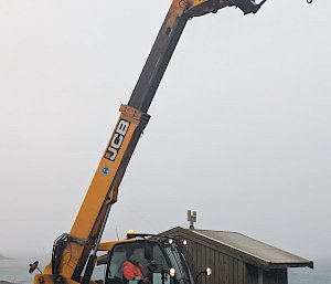 A man in an orange jacket sits in the driver's seat of a large yellow JCB telehandler. The bucket is raised high in the air.