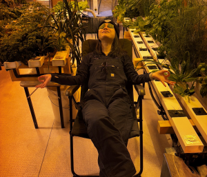 A woman sitting on a chair inside next to the station's hydroponics set up