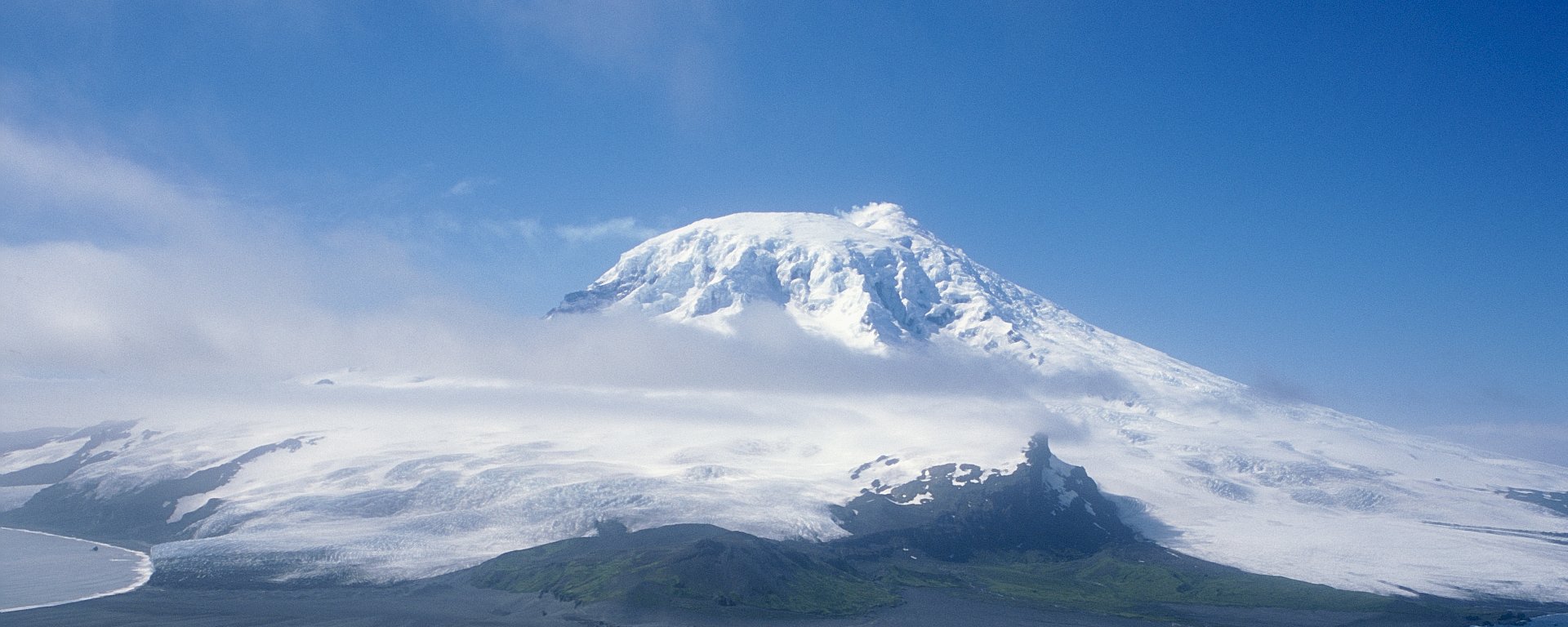 A snow covered mountain rises in the distance, with cloud around the peak and water in the foreground.