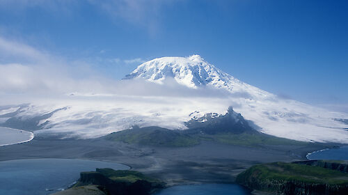A snow covered mountain rises in the distance, with cloud around the peak and water in the foreground.