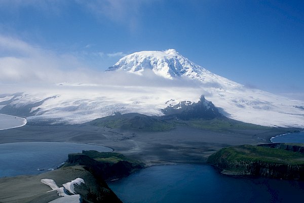 A snow covered mountain rises in the distance, with cloud around the peak and water in the foreground.