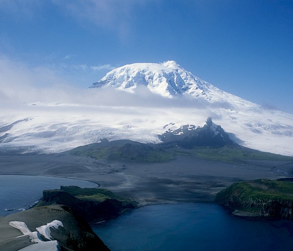 A snow covered mountain rises in the distance, with cloud around the peak and water in the foreground.