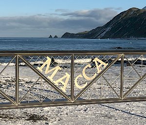 A wide silver metal gate with the words ‘Macca’ emblazoned in the centre. Looking through the gate you can see snow in the foreground, ocean in the midground and hills in the background.