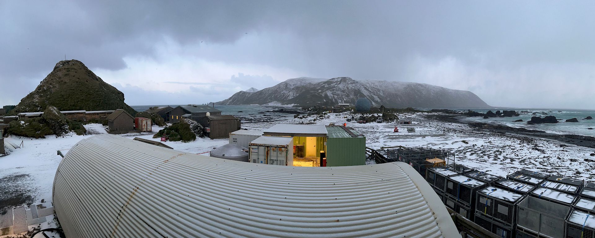 A panoramic photo of a snow-covered landscape with buildings in the foreground, ocean on either side and snow-covered mountains in the background.
