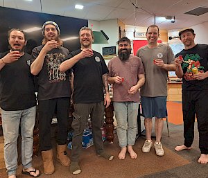 Six men stand smiling in a communal area, they are holding small glasses and all smiling.
