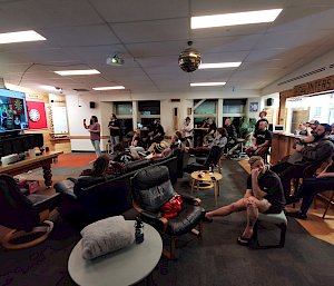 A crowd gathered around a screen and darts board watching the competition. A man is standing ready to throw a dart at the dart board.
