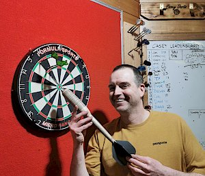 A man stands in front of a dart board with a large dart. He is smiling.