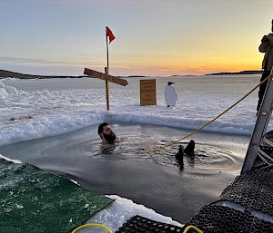 A man floating in a hole in the sea ice. He is attached to a rope and there is a cut out of a penguin on the ice next to him.