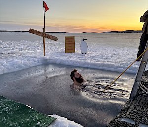A man floating in a hole in the sea ice. He is attached to a rope and there is a cut out of a penguin on the ice next to him.