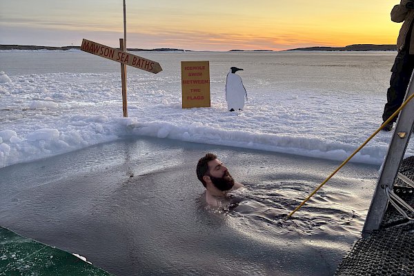 A man floating in a hole in the sea ice. He is attached to a rope and there is a cut out of a penguin on the ice next to him.