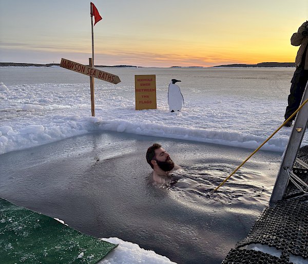 A man floating in a hole in the sea ice. He is attached to a rope and there is a cut out of a penguin on the ice next to him.