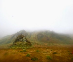 Three mist shrouded green hills in the distance with a foreground of rust coloured vegetation.