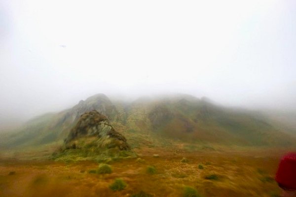 Three mist shrouded green hills in the distance with a foreground of rust coloured vegetation.