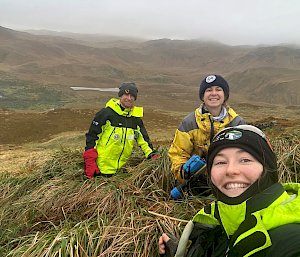 Three smiling people wearing yellow outdoor clothing stand in a misty, hilly landscape covered in tussock.