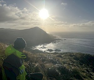A man in outdoor clothing stands atop a tussock covered hill with the sun setting in the distance over a mountain and the ocean.