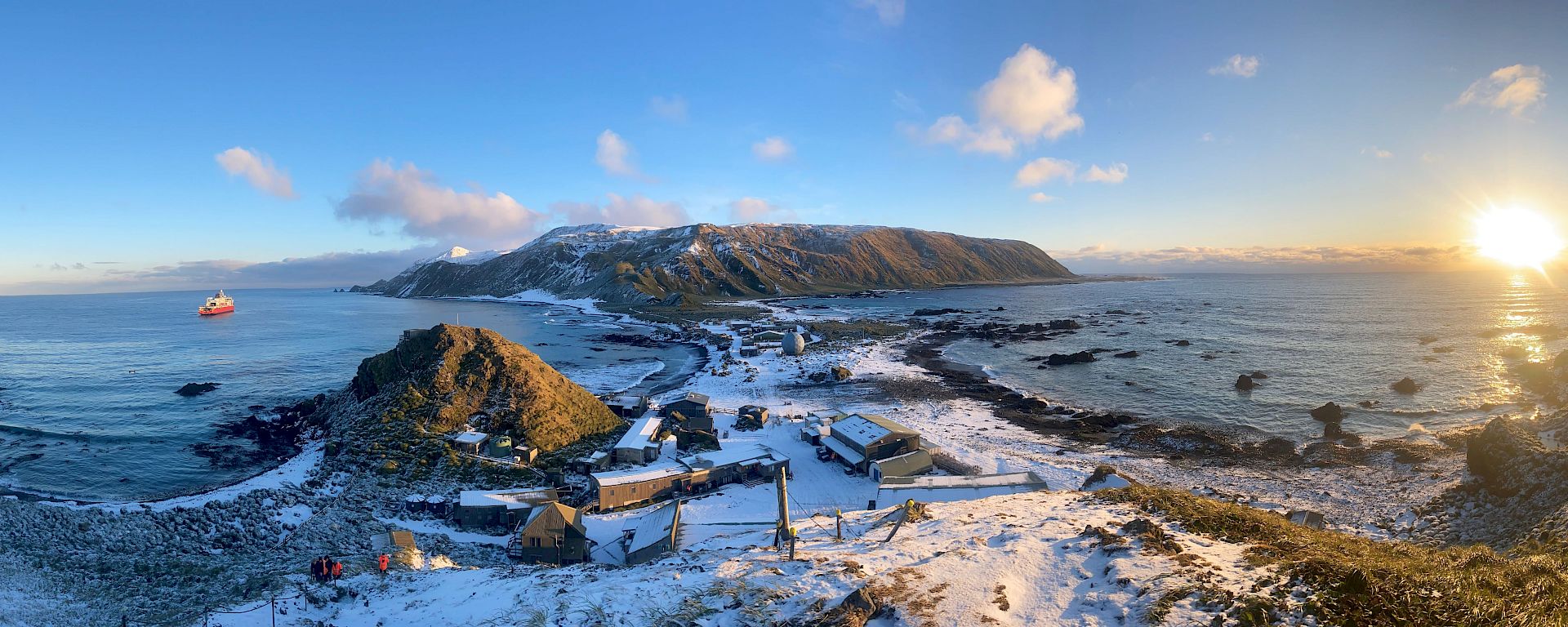 A panoramic shot of a snow-covered station and isthmus surrounded by ocean and a distant plateau covered in snow. The sun is setting in a blue sky.