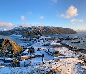 A panoramic shot of a snow-covered station and isthmus surrounded by ocean and a distant plateau covered in snow. The sun is setting in a blue sky.
