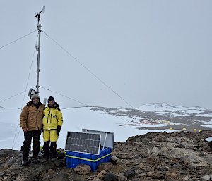 Two expeditoners standing on rocks with ice in the background next to a weather instrument on a pole