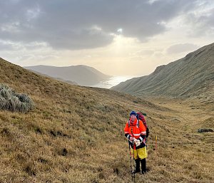 A man in an orange jacket and wearing a backpack stands in a tussock covered landscape surrounded by hills with the ocean in the distance.