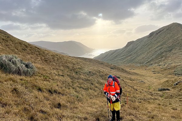 A man in an orange jacket and wearing a backpack stands in a tussock covered landscape surrounded by hills with the ocean in the distance.