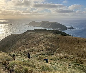 Two hikers on a track high up on a tussock covered plateau look down to an isthmus and distant headland surrounded by ocean.