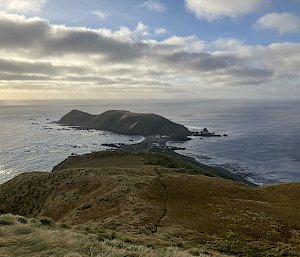 A late afternoon photo in low light looking down from a plateau towards an isthmus and a distant headland surrounded by ocean.